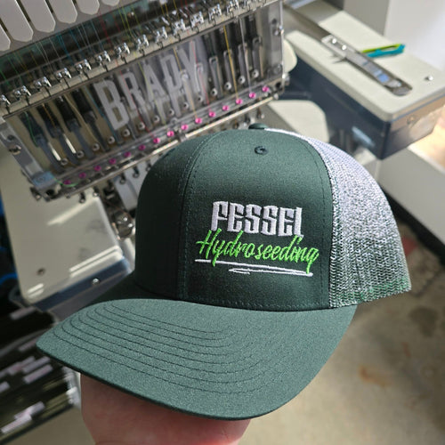 Green cap with 'Fessel Hydroseeding' text held in front of an embroidery machine.