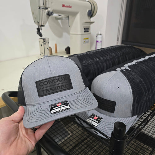Gray and black cap with branding held by a hand, with more caps in the background.