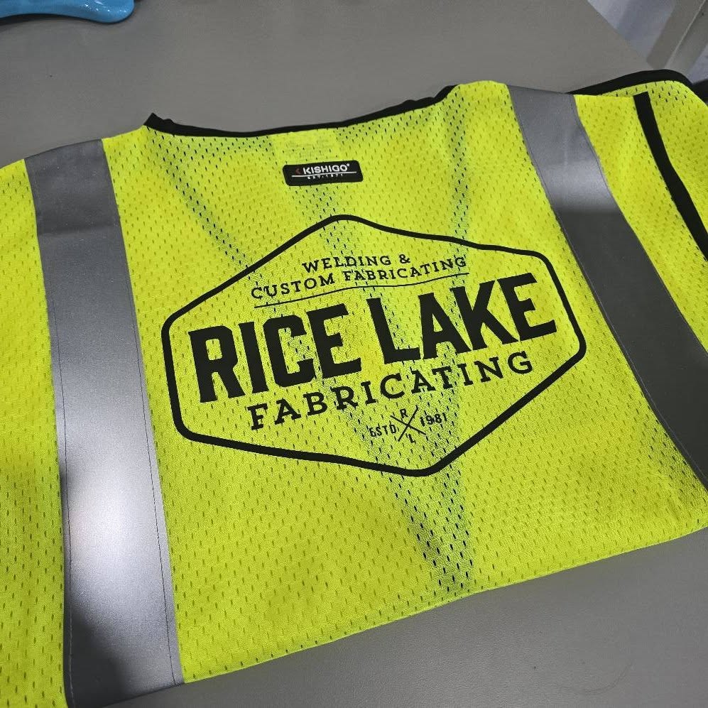 Yellow safety vest with 'Rice Lake Fabricating' logo on a gray surface